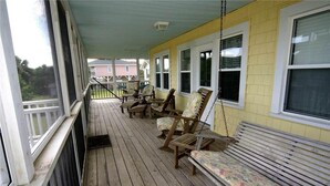 A covered porch with wooden flooring, several wooden chairs, and a bench. - Just Sew (Edisto Island)