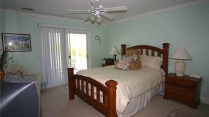 A bedroom with a wooden bed, a ceiling fan, a nightstand with a lamp, a chair, and a television. - Pink Flamingo (Edisto Island)