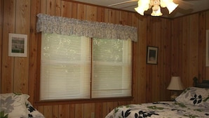 A room with wooden paneling, two beds with floral bedding, a ceiling fan, and a window with blinds.