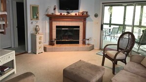 A living room with a fireplace, a television mounted above it, and a view of the outdoors through a glass door. - Summerwind 782 (Edisto Island)