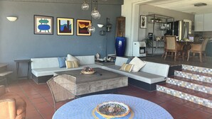 Een moderne woonkamer met een zithoek, een salontafel en een eettafel met stoelen. Er hangen kunstwerken in lijsten aan de muur, een plafondventilator en in de achtergrond is een keuken zichtbaar.