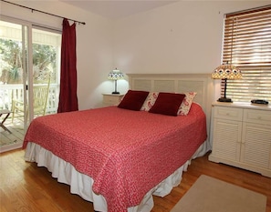 A bedroom with a large bed, a nightstand, a lamp, and a window with blinds. - Watercolors (Edisto Island)