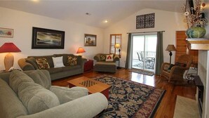 A living room with a sofa, armchairs, a coffee table, and a patterned rug. There are two lamps, a fireplace, and a view of the outside through a glass door.