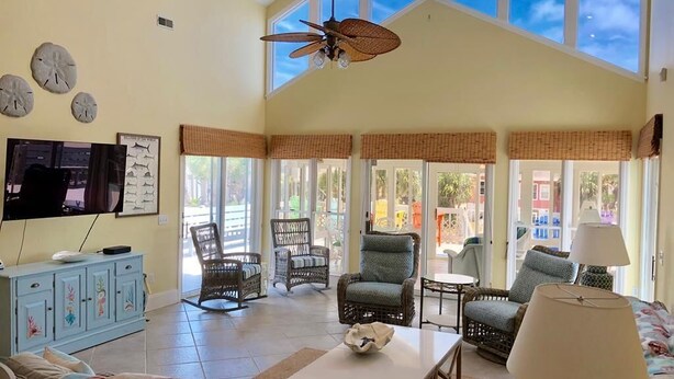 A spacious living room with a high ceiling, large windows, wicker furniture, and a television on the wall. - A Shore Investment (Edisto Island)