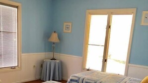 Une chambre avec deux lits, un ventilateur de plafond, une fenêtre avec des stores et une petite table avec une lampe.