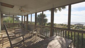 A covered outdoor deck with wooden benches and chairs, large windows, and a view of the ocean and palm trees. - Anchor inn (Edisto Island)