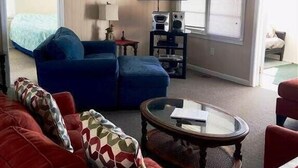 A living room with a red sofa, a blue sofa, a glass coffee table, a ceiling fan, a TV, and a window with blinds. - Beach Retreat (Edisto Island)