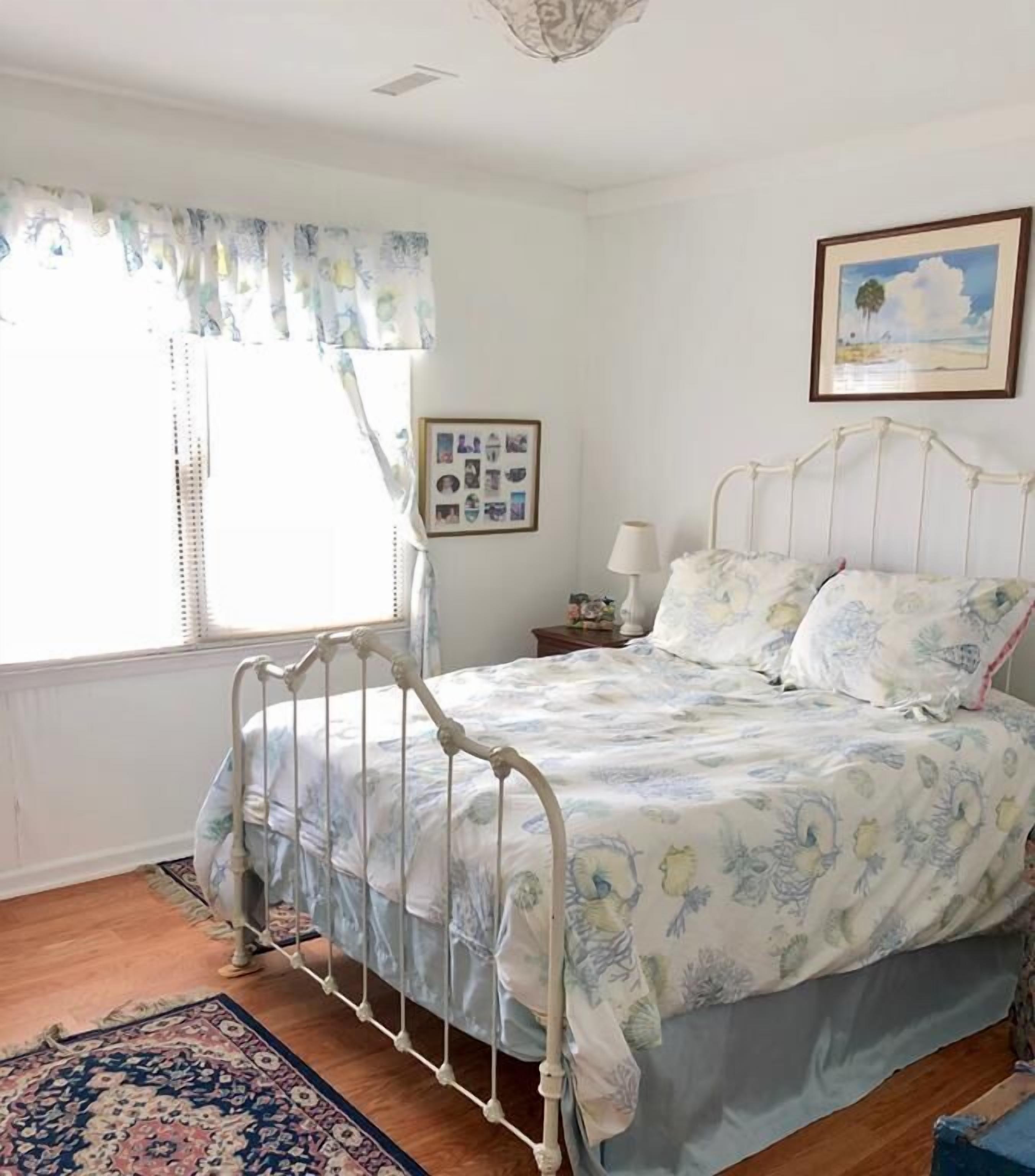 A bedroom with a metal bed frame, floral bedding, a bedside table with a lamp, a framed photo on the wall, and a window with curtains.