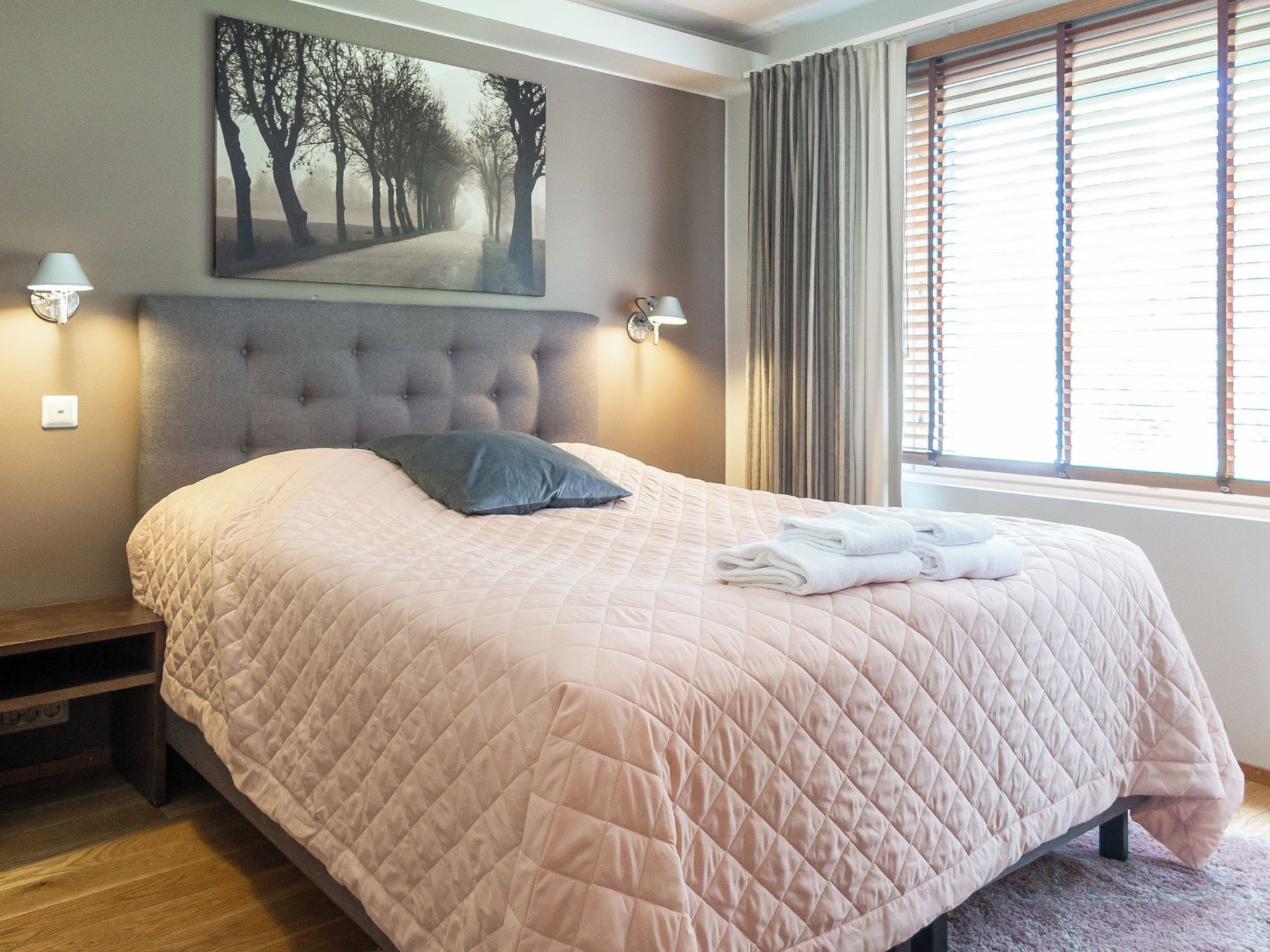 A neatly made bed with a quilted bedspread, a pillow, and folded towels on the side. There is a wall-mounted picture above the bed and two wall-mounted lamps on either side.