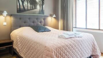 A neatly made bed with a quilted bedspread, a pillow, and folded towels on the side. There is a wall-mounted picture above the bed and two wall-mounted lamps on either side.