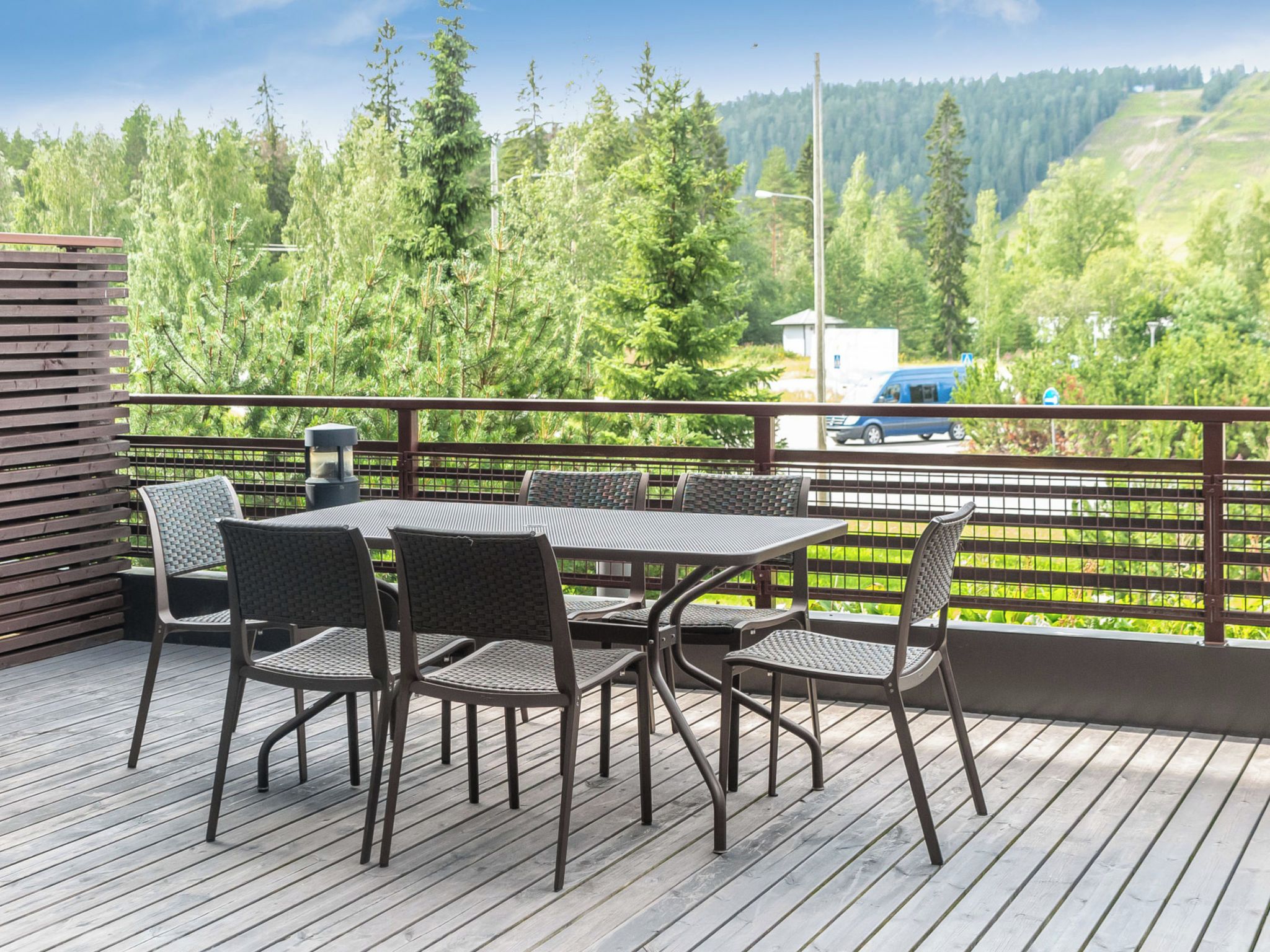 A deck with a table and chairs, overlooking a forested area with a road and vehicles in the distance.