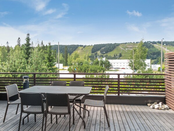 A wooden deck with a table and chairs, overlooking a green landscape with trees and buildings.