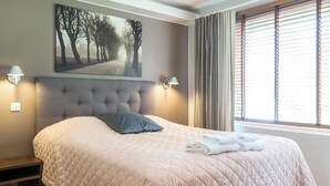 A neatly made bed with a quilted bedspread, a single pillow, and folded towels on the side. There is a wall-mounted picture of a tree-lined path, two wall-mounted lamps, and a window with blinds.