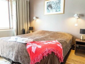 A neatly made bed with a red floral throw blanket, a pair of folded towels, and a wooden nightstand with a lamp.