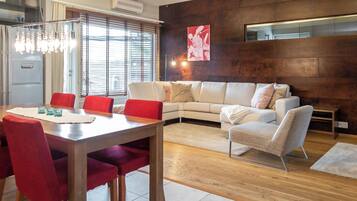 A modern living room with a dining area, featuring a sofa, chairs, a table, and a large window with blinds.