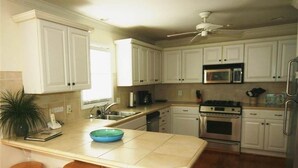Une cuisine avec des armoires blanches, un four à micro-ondes, un four et un ventilateur de plafond. On y trouve des tabourets en bois près de l’îlot central.