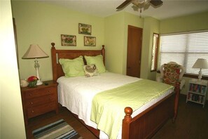 A bedroom with a wooden bed, a nightstand with a lamp, a bookshelf, and a window with blinds. - Green Parrot (Edisto Island)