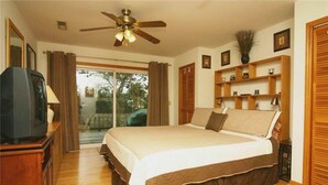 Une chambre à coucher comprenant un lit, un téléviseur, un ventilateur de plafond, une porte-fenêtre coulissante et une étagère en bois ornée d’objets décoratifs.