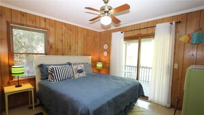 A bedroom with a bed, a ceiling fan, a window with blinds, a nightstand with a lamp, and a view of the outside through a sliding door. - Miss Irene (Edisto Island)