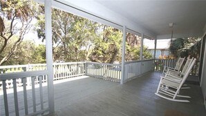 A covered porch with white railings, a rocking chair, and a view of trees and a house in the background. - Miss Irene (Edisto Island)