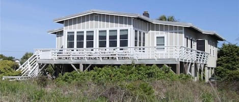 A beachfront house with a large deck, multiple windows, and a staircase leading to the entrance.