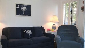 A living room with a dark blue sofa and armchair, a lamp, and a framed sign on the wall.