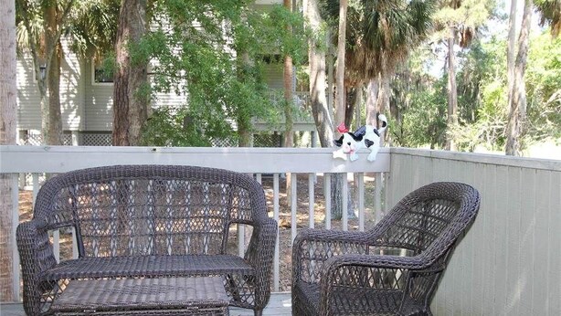 A wicker outdoor seating area with two chairs and a bench, overlooking a wooden deck and a house with a white fence.