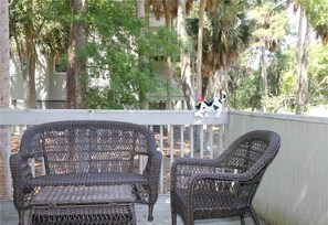 Een terras met zitgedeelte van rieten meubels, met twee stoelen en een bankje, met uitzicht op een houten terras en een huis met een witte omheining.