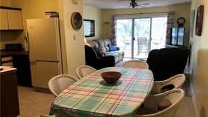 A dining area with a table set for four, surrounded by wicker chairs, a refrigerator, and a sofa in the background. - Palmetto Walk 333 (Edisto Island)