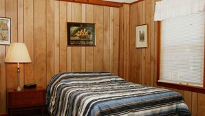 Ein Schlafzimmer mit einer Wandpaneele aus Holz, einem Bett mit gestreifter Bettdecke, einem Nachttisch mit Lampe und einem Fenster mit Jalousien.