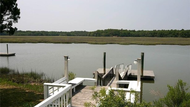 A wooden dock extending into a calm lake with a grassy shoreline and trees in the background. - Shrimpers Cove (Edisto Island)