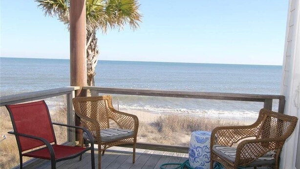 A wooden deck with two wicker chairs and a red chair, overlooking a beach and ocean. - Stormy's Newhaven (Edisto Island)