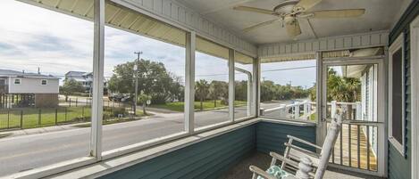 A covered porch with a ceiling fan, large windows, and a view of a street and houses.