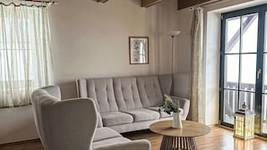 Una sala de estar con un sofá gris, una mesa redonda de madera y un ventanal con vistas a una terraza de madera.