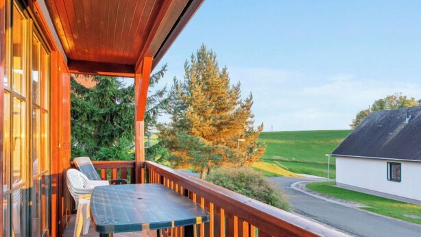 Un balcón de madera con una mesa y sillas, con vista a una carretera y campos verdes.