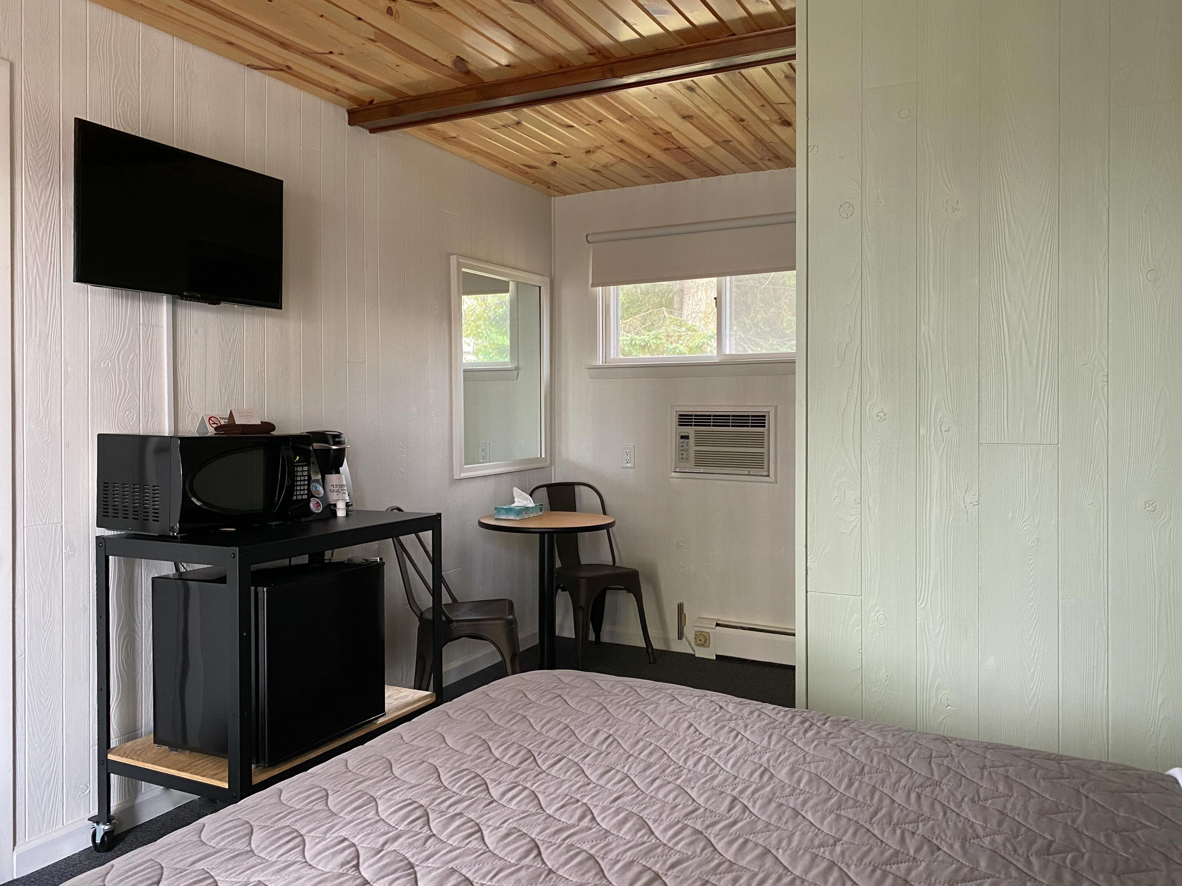 A bedroom with a bed, a desk with a microwave, a chair, a television, and a window.