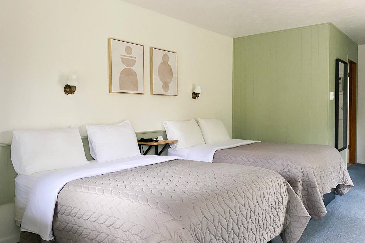 Ein Hotelzimmer mit zwei Betten, jeweils mit weißen Leinen und beigen Bettdecken. Zwei Wandleuchten und zwei gerahmte Kunstwerke befinden sich über den Betten.