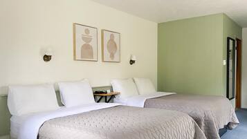 A hotel room with two beds, each with white linens and beige quilts. There are two wall-mounted light fixtures and two framed artworks above the beds.