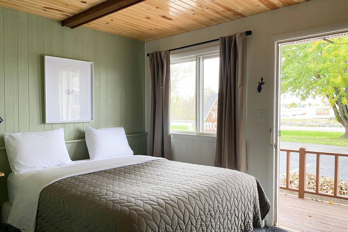 Ein Schlafzimmer mit Bett, einem Fenster mit Vorhängen, einer Tür und Blick ins Freie.