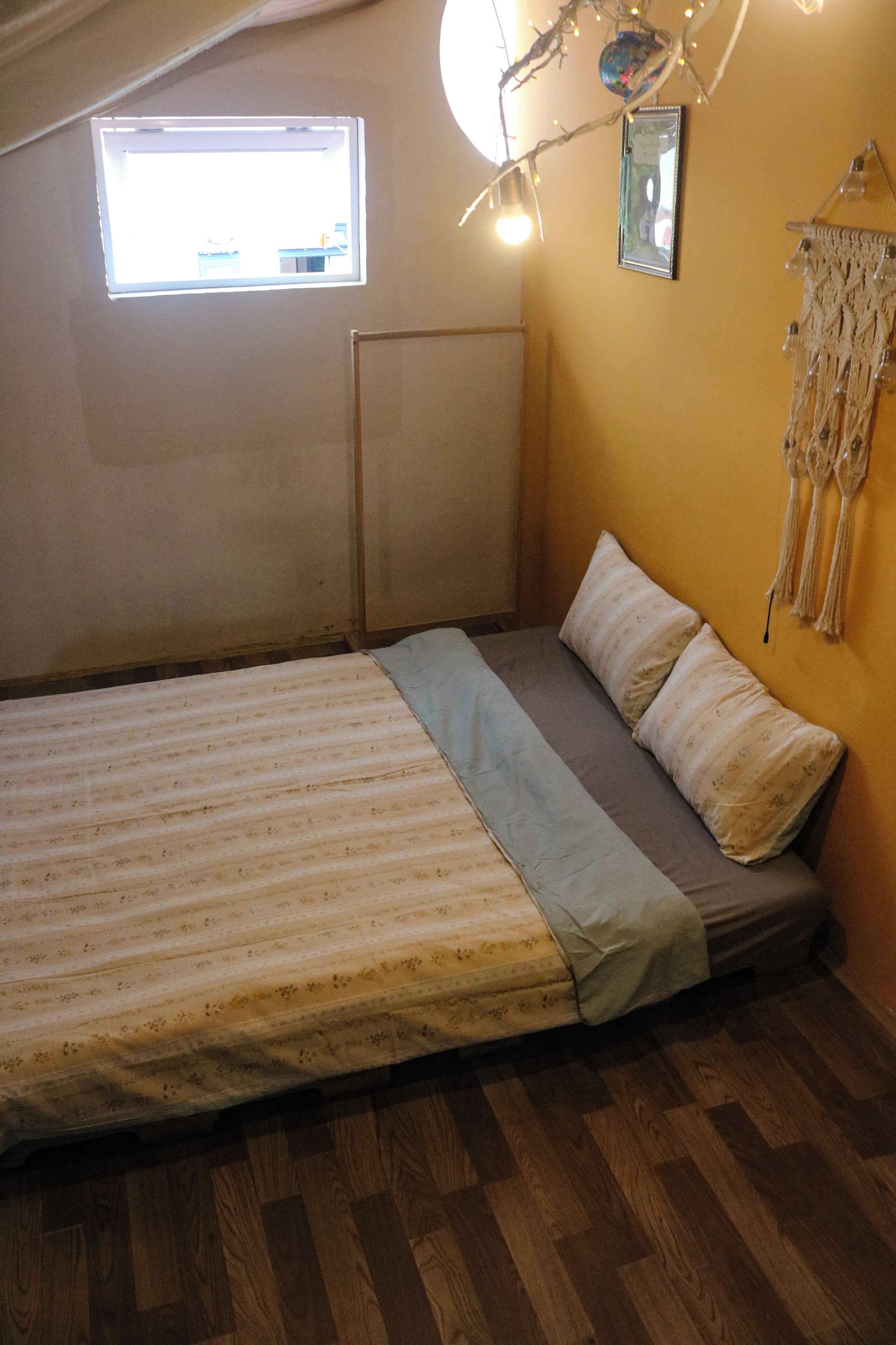A bedroom with a bed, a window, a wall-mounted picture, and a macrame wall hanging.