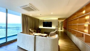 Een moderne hotelkamer met een groot raam met uitzicht op zee, een zithoek met een bank en stoelen, een wandgemonteerde flatscreen-tv en een houten vloer.