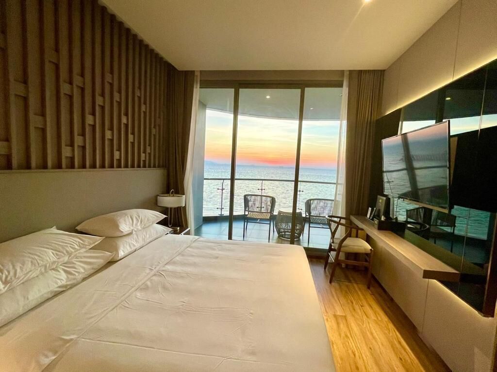 A hotel room with a large bed, a TV, a balcony with a view of the sea, and a wooden floor.