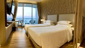 A hotel room with two beds, a TV, a desk, and a view of the sea.