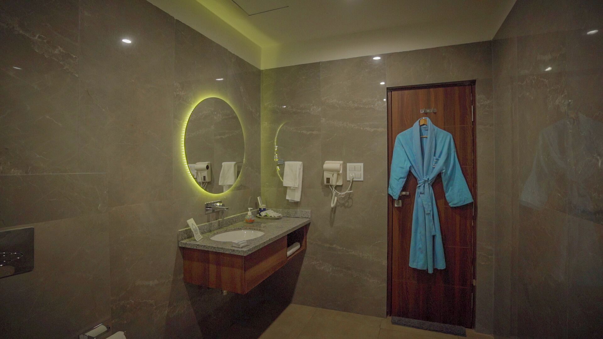 A modern bathroom with a marble wall, a wooden vanity, a sink, a mirror with a circular light, a robe hanging on the door, and a towel dispenser.