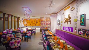 Een restaurant met een levendige inrichting, met een centraal gelegen bar, meerdere eettafels met stoelen en een decoratieve muur met culturele kunstwerken.