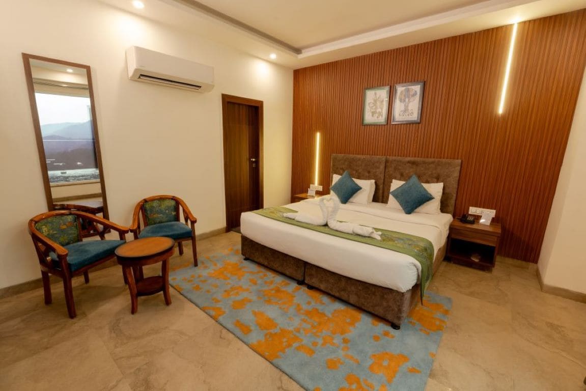 A hotel room with a bed, two chairs, a small table, an air conditioning unit, a nightstand, and a wall-mounted mirror.