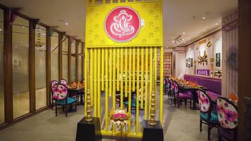 Ein Restaurant mit einer gelben Trennwand, darin ein zentrales Emblem, Tische mit farbenfroher Blumenmuster-Polsterung und einer gemusterten Wand im Hintergrund.