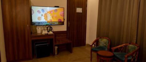 A hotel room with a television, a desk with a coffee maker, two chairs, and a bed.