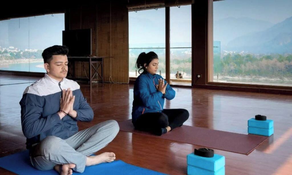 Deux personnes sont assises sur des tapis de yoga dans une pièce dotée de grandes fenêtres, de planchers en bois et offrant une vue sur un paysage.