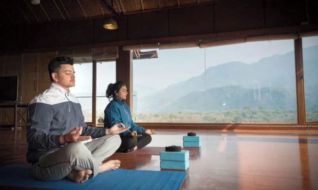 Två personer sitter i meditation på yogamattor i ett rum med stora fönster som vetter ut mot ett bergigt landskap.